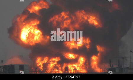 Riesige Feuer Flamme bei der Verbrennung von Öl behälter in der Raffinerie. Feuerwehrmann Wasserstrahl löschen das Feuer begann in der Nähe einer Tankstelle Lagertank. Schwarzer Regen, saurer Regen, Smog, Rauch, Asche, Umweltverschmutzung, verschwommenes Luft Stockfoto