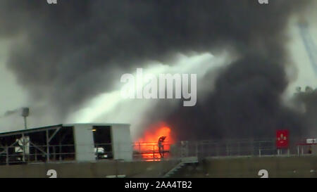 Feuerwehrmann Wasserstrahl löschen das Feuer begann in der Nähe einer Tankstelle Lagertank. Riesige Feuer Flamme bei der Verbrennung von Öl behälter in der Raffinerie. Schwarzer Regen, saurer Regen, Smog, Rauch, Asche, Umweltverschmutzung, verschwommenes Luft Stockfoto