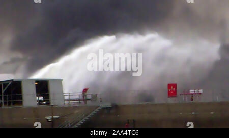 Feuerwehrmann Wasserstrahl löschen das Feuer begann in der Nähe einer Tankstelle Lagertank. Riesige Feuer Flamme bei der Verbrennung von Öl behälter in der Raffinerie. Schwarzer Regen, saurer Regen, Smog, Rauch, Asche, Umweltverschmutzung, verschwommenes Luft Stockfoto