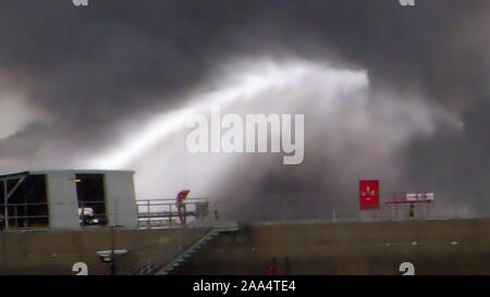 Feuerwehrmann Wasserstrahl löschen das Feuer begann in der Nähe einer Tankstelle Lagertank. Riesige Feuer Flamme bei der Verbrennung von Öl behälter in der Raffinerie. Schwarzer Regen, saurer Regen, Smog, Rauch, Asche, Umweltverschmutzung, verschwommenes Luft Stockfoto