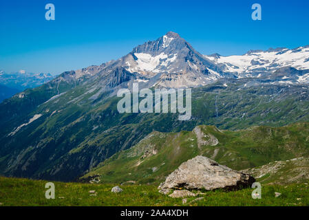 Im Sommer eine malerische Landschaft der Französischen Alpen, ein Berg mit Schnee in den Highlands, steile Hänge mit grünem Gras, ein großer Stein im Vordergrund. Stockfoto