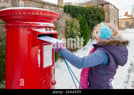 Junge Mädchen in einem schneebedeckten Szene Buchung ihr Schreiben an Santa in einem traditionellen britischen roten Briefkasten Stockfoto