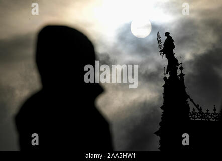 Hamburg, Deutschland. Nov, 2019 19. Ein Mann schaut auf die Statue auf einem Giebel der Hamburger Rathaus, die von der Sonne durch dunkle Wolken geglänzt. Quelle: Axel Heimken/dpa/Alamy leben Nachrichten Stockfoto