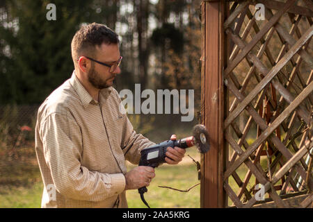 Mann mit Brille und ein flanellhemd im Frühling im Garten arbeiten. Renovierung und Instandhaltung von Garten Pergolen. Entfernen alter Farbe von Elementen aus Holz. Stockfoto