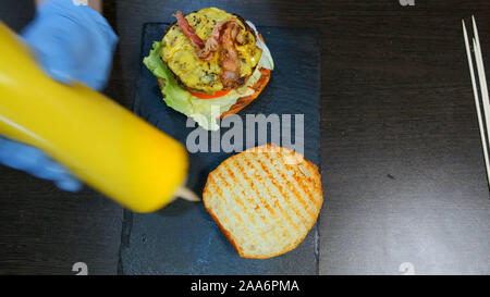 Close-up Blick von oben auf die Hände, ein Koch, der einen Hamburger. Sauce auf einem Brötchen und eine leckere und schmackhafte Burger zusammenbauen Stockfoto