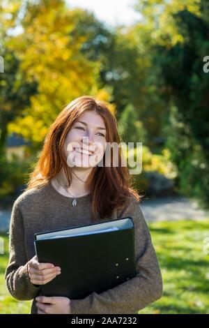 Junge lächelnde rothaarige Frau, Student, Schüler hält Ordner, Bayern, Deutschland Stockfoto