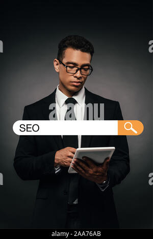 Afrikanische amerikanische Geschäftsmann in Gläsern mit digitalen Tablet auf dunklem Hintergrund mit Seo-Schriftzug auf der Suche nach Bar Abbildung Stockfoto