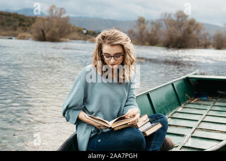 Porträt der jungen schönen Frau, die auf einem Boot sitzt und ein Buch liest. Sie ist Bücherwurm und sie wählt zwischen wenigen Büchern. Stockfoto