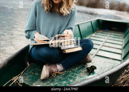 Porträt der jungen schönen Frau, die auf einem Boot sitzt und ein Buch liest. Sie ist Bücherwurm und sie wählt zwischen wenigen Büchern. Stockfoto