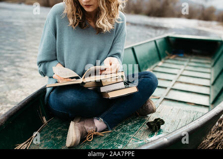 Porträt der jungen schönen Frau, die auf einem Boot sitzt und ein Buch liest. Sie ist Bücherwurm und sie wählt zwischen wenigen Büchern. Stockfoto