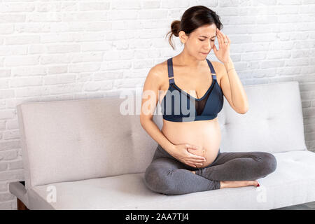 Müde Asiatischen schwangere Frau berühren Kopf und Magen leiden unter Kopfschmerzen auf dem Sofa sitzen mit gekreuzten Beinen Stockfoto