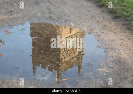 Spiegelung des britischen Kirchturms mit der Kirchenuhr, die nach heftigen Regenfällen kopfüber in einer Pfütze auf dem Boden eines schlammigen Pfades gesehen wird. Stockfoto