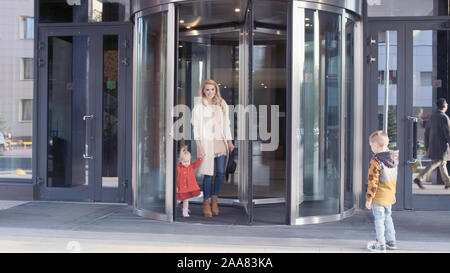 Reisende mit Familie verlassen das Hotel durch Glastüren. Mutter mit Kindern gehen Stockfoto