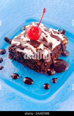 Hausgemachte Schokolade Fudge Brownie garniert mit Sahne und eine maraschino Kirsche. Auf einem bunten blauen Platte serviert. Stockfoto