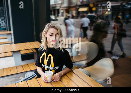 Schöne junge Frau vor McDonald's in Sarajewo sitzen. Sie ist trinken Soda und das Tragen von McDonald's T-Shirt. August, 2019. Stockfoto