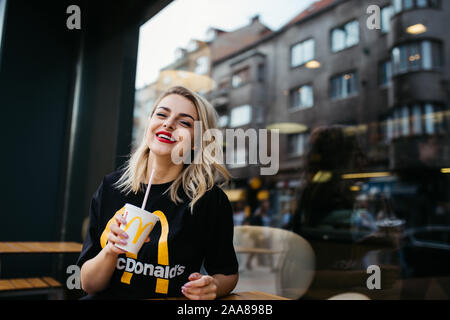 Schöne junge Frau vor McDonald's in Sarajewo sitzen. Sie ist trinken Soda und das Tragen von McDonald's T-Shirt. August, 2019. Stockfoto