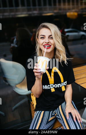 Schöne junge Frau vor McDonald's in Sarajewo sitzen. Sie ist trinken Soda und das Tragen von McDonald's T-Shirt. August, 2019. Stockfoto