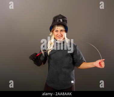 Junge blonde Mädchen in einem fechten Trainer dunklen Uniform und schwarzen Helm Maske steht mit einem rapier auf grauem Hintergrund Stockfoto