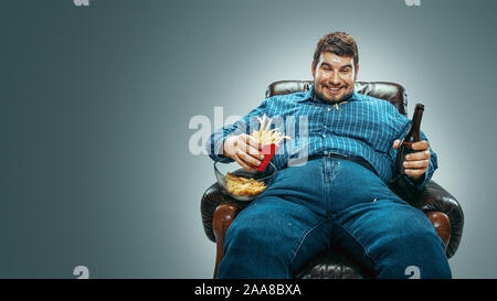 Portrait von Fett kaukasischen Mann und jeanse whirt in einem braunen Sessel auf Farbübergang grauer Hintergrund sitzen. Fernsehen trinkt Bier, isst Chips, gebratene Kartoffeln, Laughting. Übergewicht, sorglos. Stockfoto