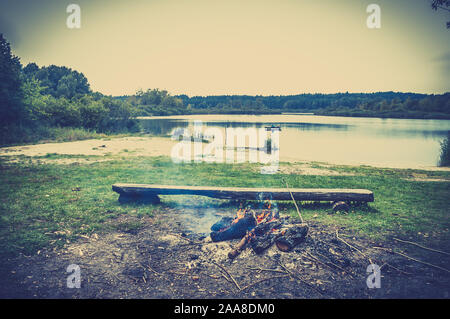 Schöne Landschaft von Lagerfeuer am See in der Nähe des Waldes. Boot mit Fischer auf dem See im Hintergrund, vintage Foto. Stockfoto