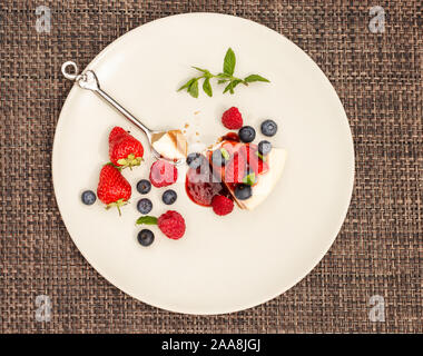 Käsekuchen mit Erdbeeren, Heidelbeeren, Himbeeren, Obst Coulis und Minze auf eine Creme Platte mit braunen Unterlage Hintergrund von oben Stockfoto