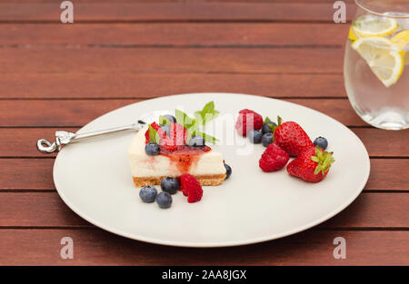 Käsekuchen mit Früchten, Beeren, Coulis und Minze mit Glas Eiswasser und Zitrone auf hölzernen Tisch Stockfoto