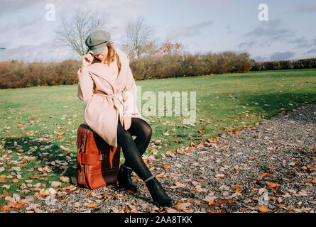 Frau sitzt auf einem Koffer warten auf einer Landstraße im Herbst Stockfoto