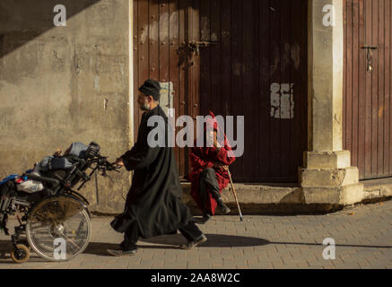 Szenen des täglichen Lebens in der Medina von Fes. Alter Mann Sonnenbaden inzwischen ein Vater seinem Kind tetraplegic tragen. Stockfoto