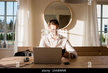 Mann bei der Arbeit von zu Hause aus auf sein Handy und Laptop konzentrieren. Stockfoto