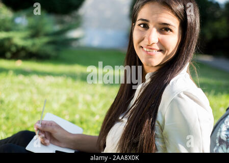 Porträt eines jungen indischen Studenten, Geschäftsfrau. Eine Frau mit einem Rucksack in einem Rucksack hält eine Tablette, sitzt auf dem grünen Gras, gegen die Sonne. Su Stockfoto