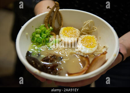 Frau Hände halten eine Schüssel mit traditionellen Japanischen Schweinefleisch Ramen Suppe, mit enoki Pilze, mittlere gekochte Eier, Schalotten, Algen und Schweinefleisch Stockfoto
