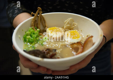 Frau Hände halten eine Schüssel mit traditionellen Japanischen Schweinefleisch Ramen Suppe, mit enoki Pilze, mittlere gekochte Eier, Schalotten, Algen und Schweinefleisch Stockfoto