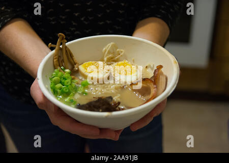 Frau Hände halten eine Schüssel mit traditionellen Japanischen Schweinefleisch Ramen Suppe, mit enoki Pilze, mittlere gekochte Eier, Schalotten, Algen und Schweinefleisch Stockfoto