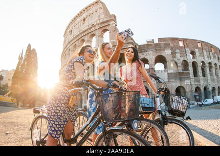 Drei glückliche junge Frauen Freunde Touristen mit Fahrrädern unter selfies am Kolosseum in Rom, Italien bei Sonnenaufgang. Stockfoto