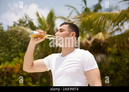 Junger Mann Bier trinken am tropischen Strand mit Kokospalmen auf Hintergrund. Kaukasier Männlich mit einer Flasche Limonade trinken stillt Durst Stockfoto