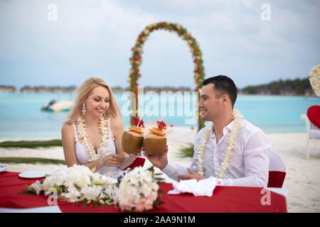 Bräutigam und Braut drink coconut Cocktails an der Hochzeit Tisch mit lei von Blumen auf den Strand von tropischen Insel auf den Malediven. Türkisfarbene Meer und die Zeremonie Stockfoto