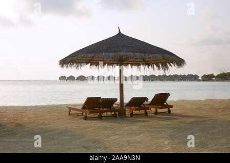 Strand Liegestühle unter Sonnenschirmen Stroh. Küste des Indischen Ozeans auf den Malediven Insel. Weißer Sandstrand und ruhige See. Reisen und Ferien. Stockfoto