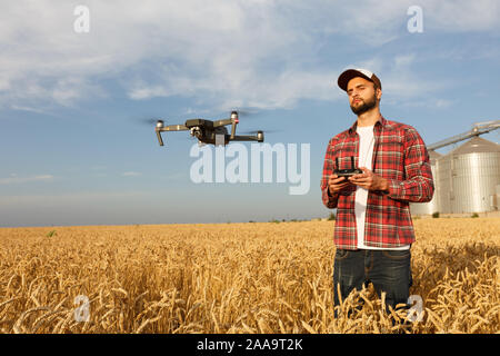 Drone schwebt vor Bauer mit Fernbedienung in der Hand in der Nähe des Kornelevators. Quadcopter fliegt in der Nähe von Pilot. Agrarwissenschaftler, Luftaufnahmen und Stockfoto