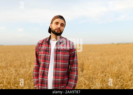 Portrait eines bärtigen Landwirt in einem Weizenfeld. Stylisches hipster Mann mit Trucker hat und kariertem Hemd auf. Landwirtschaftliche Arbeitnehmer Stockfoto