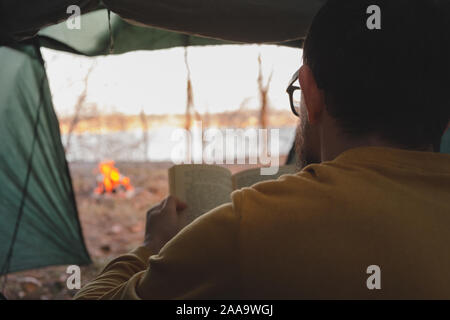 Mann sitzt im Zelt am Lagerfeuer und liest ein Buch. Genießen Sie die ruhige erholsame Ferien in der Natur, das Konzept der Ruhe und weg von Urb. Stockfoto