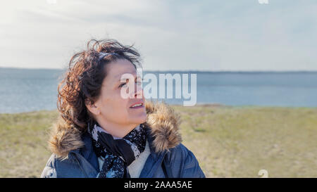 Eine Frau mittleren Alters an der Küste in casual Winter Kleidung sieht die Seite voller Optimismus, Hoffnung und Glück. Kopieren Sie Platz an der Seite. Stockfoto