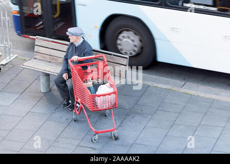 Ein älterer weißer Mann Mann sitzt auf einer Bank, Sitz in der Nähe einer Bushaltestelle Holding einen Einkaufswagen und Suchen auf der Straße als Bus verläuft in der Nähe Stockfoto