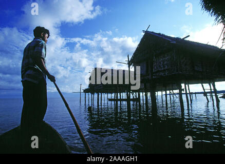 Indonesien. Sulawesi. Bajo Mann stand im Kanu durch Waterside pole Häuser. Stockfoto