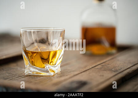 Nahaufnahme von einem Glas mit golden schimmernden Scotch Whisky auf einem alten rustikalen Holzmöbeln, unscharfe Whisky Flasche im Hintergrund Stockfoto