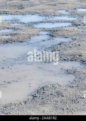 Sonnenschein auf einem kaputten Hardcore-Parkplatz mit Pfützen stehendem Wasser in Hohlräumen/Schlaglochen. Blauer Himmel, reflektiert in Pfützen, Wasserreflexionen. Stockfoto