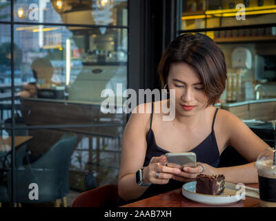 Schöne happy asiatische Frau kurze Haare mit Smartphone und trinken Kaffee und Dessert im Cafe. Stockfoto