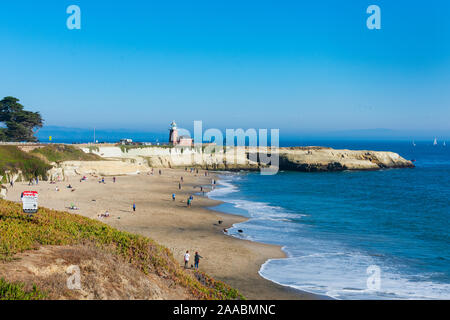 Bewohner, Besucher, Touristen genießen das Wasser des Pazifischen Ozeans auf sandigen Hund Strand. Marke Abbott Memorial Leuchtturm im Hintergrund. - Santa Cruz, Kalifornien, USA - 2019 Stockfoto