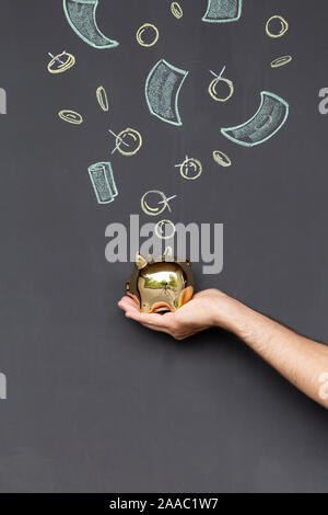 Konzept der Geld sparen mit einem goldenen Sparschwein in der Hand vor einer Tafel mit Hand gezeichnet Münzen und Banknoten gehalten Stockfoto