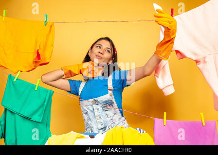 Lustige und schöne Hausfrau Hausarbeit tun auf gelbem Hintergrund. Junge kaukasier Frau durch gewaschene Kleidung umgeben. Das häusliche Leben, helle Artwork, allgemeine Ordnung und Sauberkeit. Die selfie. Stockfoto