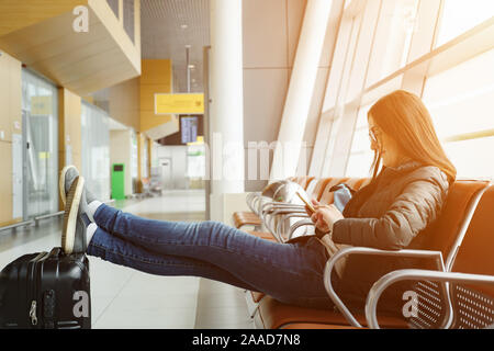 Junge brünette Frau in Flughafen wartet auf Ihren Flug, etwas am Smartphone suchen. Sie sitzt mit ihrer Beine ausgestreckt und setzt sie auf den Koffer, Sonnenlicht Stockfoto